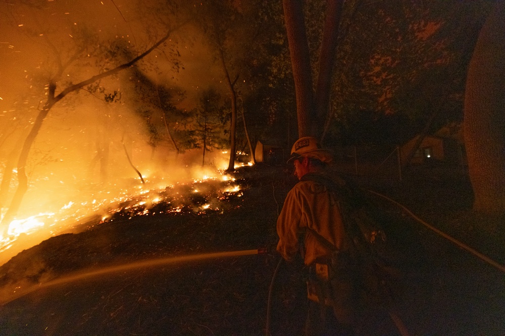 A firefighter is battling a wildfire at night in a forested area, directing a water hose toward advancing flames on a hillside. The fire is spreading along the ground and through the trees, producing intense orange glow, embers, and smoke. A structure appears faintly visible in the background to the right.