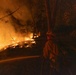 A firefighter is battling a wildfire at night in a forested area, directing a water hose toward advancing flames on a hillside. The fire is spreading along the ground and through the trees, producing intense orange glow, embers, and smoke. A structure appears faintly visible in the background to the right.