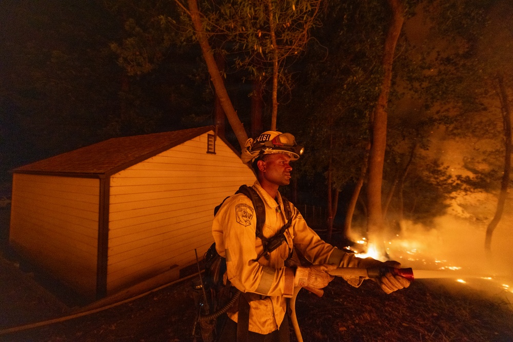 A firefighter in full protective gear holds a fire hose while battling an active wildfire at night. Flames are visible in the background among trees, with a small outbuilding or shed nearby that appears to be at risk. The scene is dramatically lit by the orange glow of the fire.