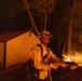 A firefighter in full protective gear holds a fire hose while battling an active wildfire at night. Flames are visible in the background among trees, with a small outbuilding or shed nearby that appears to be at risk. The scene is dramatically lit by the orange glow of the fire.
