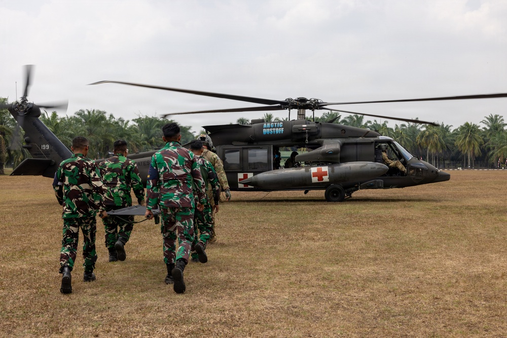 Military personnel in camouflage uniforms carry a stretcher toward a UH-60 Black Hawk medical evacuation helicopter marked with Red Cross symbols and labeled 'Arctic Dustoff'. The scene appears to be a MEDEVAC training exercise or operation conducted on an open field surrounded by tropical palm trees. Personnel from what appear to be two different military forces — one in Indonesian TNI-pattern camouflage and others in US Army uniforms — are working together.