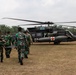 Military personnel in camouflage uniforms carry a stretcher toward a UH-60 Black Hawk medical evacuation helicopter marked with Red Cross symbols and labeled 'Arctic Dustoff'. The scene appears to be a MEDEVAC training exercise or operation conducted on an open field surrounded by tropical palm trees. Personnel from what appear to be two different military forces — one in Indonesian TNI-pattern camouflage and others in US Army uniforms — are working together.