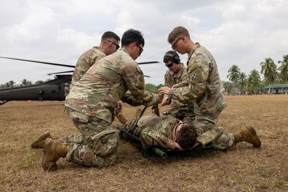 Four U.S. Army soldiers in OCP camouflage uniforms are conducting a medical casualty evacuation (CASEVAC) training exercise in a field. They are attending to a simulated casualty on a litter/stretcher, with a United States Army UH-60 Black Hawk helicopter visible in the background. The setting appears to be a tropical location based on the palm trees and vegetation.
