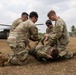 Four U.S. Army soldiers in OCP camouflage uniforms are conducting a medical casualty evacuation (CASEVAC) training exercise in a field. They are attending to a simulated casualty on a litter/stretcher, with a United States Army UH-60 Black Hawk helicopter visible in the background. The setting appears to be a tropical location based on the palm trees and vegetation.
