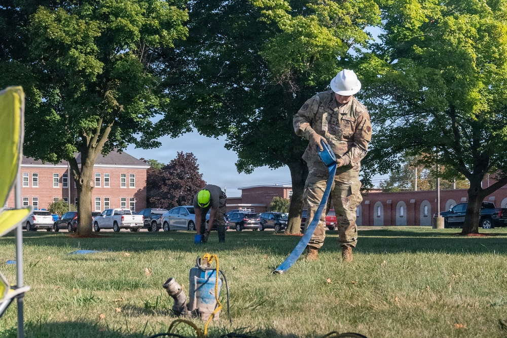 Two workers are performing what appears to be water pumping or drainage operations on a grassy area, likely on a military installation or institutional campus. One worker in military camouflage uniform and white hard hat is handling a blue hose connected to a pump, while a second worker in civilian clothes with a green hard hat is bent over working in the background. Brick institutional buildings and a parking lot with vehicles are visible in the background.