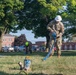 Two workers are performing what appears to be water pumping or drainage operations on a grassy area, likely on a military installation or institutional campus. One worker in military camouflage uniform and white hard hat is handling a blue hose connected to a pump, while a second worker in civilian clothes with a green hard hat is bent over working in the background. Brick institutional buildings and a parking lot with vehicles are visible in the background.