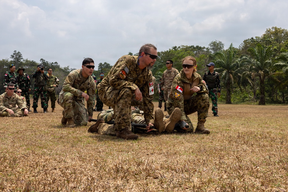 Military personnel from multiple nations are conducting a field medical training exercise on an open grassy area. Two individuals wearing Canadian Armed Forces patches with red cross medical insignia are demonstrating casualty care techniques on a simulated patient on the ground. A multinational audience including soldiers in Indonesian camouflage uniforms observes the demonstration in the background.