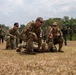 Military personnel from multiple nations are conducting a field medical training exercise on an open grassy area. Two individuals wearing Canadian Armed Forces patches with red cross medical insignia are demonstrating casualty care techniques on a simulated patient on the ground. A multinational audience including soldiers in Indonesian camouflage uniforms observes the demonstration in the background.