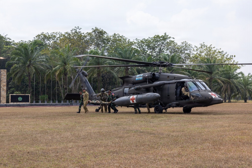 A military medical evacuation (MEDEVAC) exercise is taking place on an open field surrounded by tropical vegetation, including palm trees. Personnel in mixed military uniforms are loading or unloading a patient on a stretcher into/from a dark-colored UH-60 Black Hawk helicopter marked with Red Cross insignia. The helicopter is labeled 'ARCTIC DUSTOFF,' indicating a U.S. Army MEDEVAC unit, and appears to be conducting a joint exercise with Indonesian military personnel.