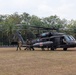 A military medical evacuation (MEDEVAC) exercise is taking place on an open field surrounded by tropical vegetation, including palm trees. Personnel in mixed military uniforms are loading or unloading a patient on a stretcher into/from a dark-colored UH-60 Black Hawk helicopter marked with Red Cross insignia. The helicopter is labeled 'ARCTIC DUSTOFF,' indicating a U.S. Army MEDEVAC unit, and appears to be conducting a joint exercise with Indonesian military personnel.