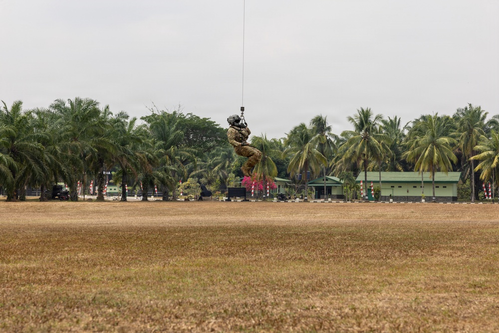 A military personnel in full combat gear is suspended mid-air from a cable or rope, likely being hoisted by a helicopter above a dry, open field. The setting appears to be a military installation or training facility in a tropical region, identifiable by the palm trees and green buildings in the background. This appears to be a helicopter hoist or SPIE (Special Purpose Insertion/Extraction) training exercise.
