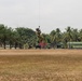 A military personnel in full combat gear is suspended mid-air from a cable or rope, likely being hoisted by a helicopter above a dry, open field. The setting appears to be a military installation or training facility in a tropical region, identifiable by the palm trees and green buildings in the background. This appears to be a helicopter hoist or SPIE (Special Purpose Insertion/Extraction) training exercise.