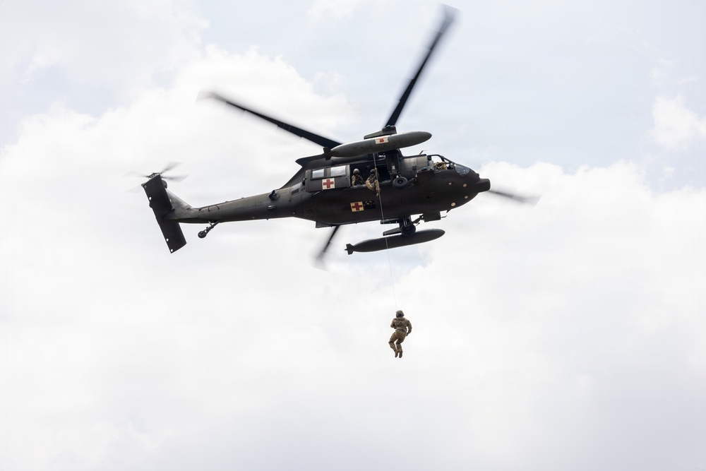 A dark military helicopter, identifiable as a medevac variant by the red cross markings on its fuselage, is hovering in a partly cloudy sky. A soldier in combat gear is suspended below the aircraft on a hoist or rope, indicating a medical evacuation or rescue training exercise. The image is captured from a low angle looking upward, providing a clear view of the aircraft's underside and the suspended personnel.