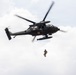A dark military helicopter, identifiable as a medevac variant by the red cross markings on its fuselage, is hovering in a partly cloudy sky. A soldier in combat gear is suspended below the aircraft on a hoist or rope, indicating a medical evacuation or rescue training exercise. The image is captured from a low angle looking upward, providing a clear view of the aircraft's underside and the suspended personnel.