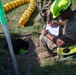 A military or rescue personnel wearing a PMI-branded high-visibility green helmet sits in a camp chair beside an open underground access hole, writing on a clipboard. Safety equipment including chains as a perimeter barrier, a ventilation duct (yellow flexible tubing), and a metal support structure are visible. A second helmeted person is partially visible in the background, and a green helmet can be seen inside the hole, suggesting an active confined space entry operation.