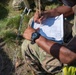 A soldier in MultiCam/OCP camouflage uniform is writing on a clipboard with a pencil while seated outdoors on grass. The individual is wearing a black G-Shock watch and has a visible tattoo on their forearm. The scene appears to be a field training exercise or operational environment.