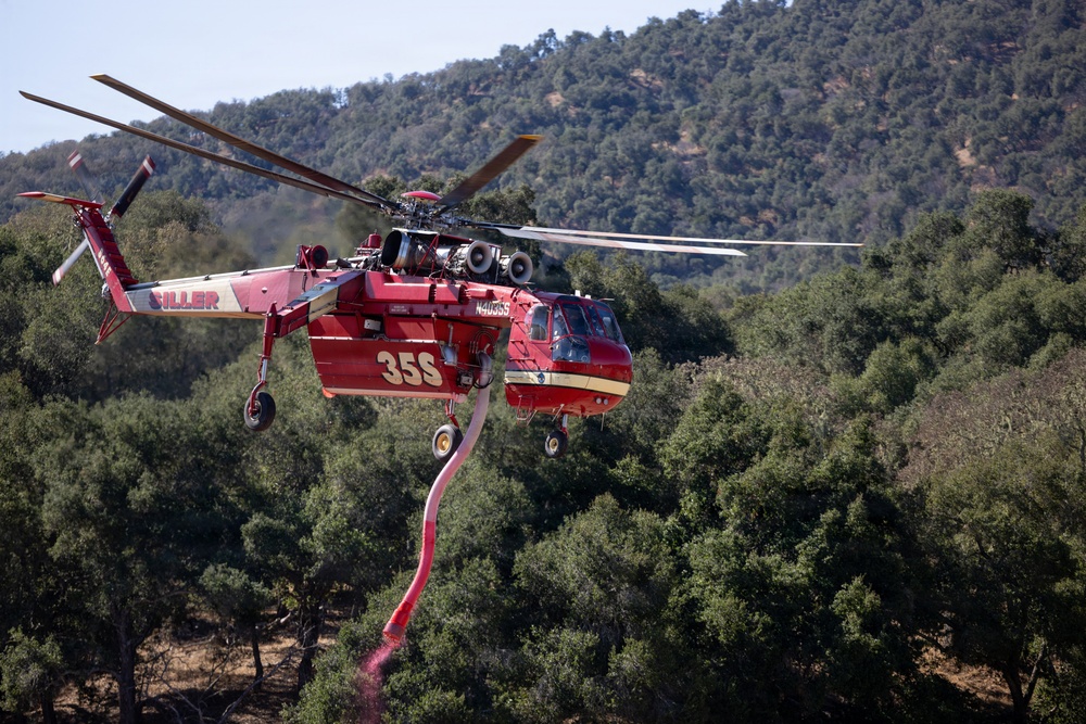 A red firefighting helicopter identified as 'Siller' with tail number N4036S and designation '35S' is captured in flight over a densely forested hillside. The helicopter, consistent with a Sikorsky S-64 Aircrane or similar heavy-lift type, is actively dispensing what appears to be red fire retardant from a hose beneath its belly tank. The background shows a green, tree-covered mountain slope under clear skies.