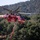 A red firefighting helicopter identified as 'Siller' with tail number N4036S and designation '35S' is captured in flight over a densely forested hillside. The helicopter, consistent with a Sikorsky S-64 Aircrane or similar heavy-lift type, is actively dispensing what appears to be red fire retardant from a hose beneath its belly tank. The background shows a green, tree-covered mountain slope under clear skies.