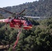 A red firefighting helicopter identified as 'Siller' with tail number N4036S and designation '35S' is captured in flight over a densely forested hillside. The helicopter, consistent with a Sikorsky S-64 Aircrane or similar heavy-lift type, is actively dispensing what appears to be red fire retardant from a hose beneath its belly tank. The background shows a green, tree-covered mountain slope under clear skies.
