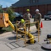Military or utility personnel are conducting a confined space entry operation at an open manhole on a paved surface near a brick building. A ventilation duct (yellow flexible hose) runs from an orange blower/ventilation unit into the manhole, with a safety tripod/guardrail system erected around the opening. Three individuals are present — two wearing green helmets and one in military camouflage with a white hard hat — overseeing or documenting the operation.