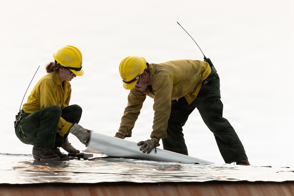 Two wildland firefighters wearing yellow hard hats marked 'E-317' are working together on what appears to be a reflective fire shelter or aluminum foil-like protective material on a surface. They are deploying or rolling out a cylindrical object, likely a hose or rolled material, on top of the reflective sheeting. Both workers are equipped with radios and wearing standard wildland firefighting gear.