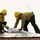 Two wildland firefighters wearing yellow hard hats marked 'E-317' are working together on what appears to be a reflective fire shelter or aluminum foil-like protective material on a surface. They are deploying or rolling out a cylindrical object, likely a hose or rolled material, on top of the reflective sheeting. Both workers are equipped with radios and wearing standard wildland firefighting gear.
