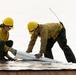 Two wildland firefighters wearing yellow hard hats marked 'E-317' are working together on what appears to be a reflective fire shelter or aluminum foil-like protective material on a surface. They are deploying or rolling out a cylindrical object, likely a hose or rolled material, on top of the reflective sheeting. Both workers are equipped with radios and wearing standard wildland firefighting gear.