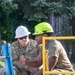 Two U.S. Air Force personnel in OCP (Operational Camouflage Pattern) uniforms are crouched down reviewing documents on the ground near construction equipment. The individual on the left, identified by name tape as 'WOOTEN,' wears a white hard hat and sunglasses, while the individual on the right wears a bright green 'PMI' branded hard hat. They appear to be conducting a safety briefing or planning discussion at a work site.
