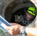 A worker wearing a bright green safety helmet with a mounted headlamp and a safety harness is positioned inside a circular concrete manhole or tunnel opening. They are reaching upward with a gloved hand toward another person above ground who is holding what appears to be a clipboard or document. The scene depicts a confined space entry operation, likely an inspection or maintenance task.