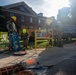Military personnel in combat uniforms and hard hats are conducting utility or infrastructure work near an open manhole on a sidewalk adjacent to a brick building. Workers are using a blue ladder, yellow safety barriers, and traffic cones to secure the work area. The scene appears to be on a military installation or government campus based on the architecture and personnel attire.