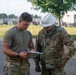 Two U.S. Air Force personnel are reviewing documents on a clipboard outdoors on what appears to be a military installation. One individual is in a green t-shirt and camouflage pants, while the other wears a full OCP uniform with a white hard hat, indicating a construction or engineering oversight role. The 'U.S. AIR FORCE' tape is clearly visible on the uniformed individual's chest.