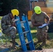 Two military personnel in OCP (Operational Camouflage Pattern) uniforms are working at what appears to be a utility access point or manhole site. One soldier wearing a yellow hard hat is seated in a folding chair reviewing documents, while the other wearing a neon green hard hat is kneeling and writing on a clipboard. A blue Werner ladder is positioned near an open manhole, suggesting confined space entry operations.