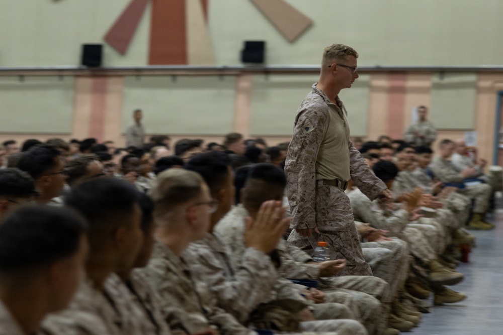 A large group of military personnel in desert camouflage uniforms are seated in a gymnasium or assembly hall. One service member is standing and walking among the seated crowd, appearing to be in a position of authority or addressing the group. The setting suggests a formal military briefing, all-hands meeting, or command assembly.