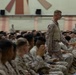 A large group of military personnel in desert camouflage uniforms are seated in a gymnasium or assembly hall. One service member is standing and walking among the seated crowd, appearing to be in a position of authority or addressing the group. The setting suggests a formal military briefing, all-hands meeting, or command assembly.