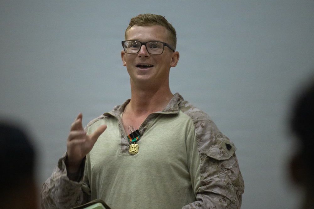 A male service member in U.S. Marine Corps MARPAT camouflage uniform is speaking or addressing an audience. He is wearing glasses and has a military medal or award hanging around his neck on a ribbon. His right hand is raised in a gesturing motion, suggesting he is actively speaking.