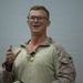 A male service member in U.S. Marine Corps MARPAT camouflage uniform is speaking or addressing an audience. He is wearing glasses and has a military medal or award hanging around his neck on a ribbon. His right hand is raised in a gesturing motion, suggesting he is actively speaking.