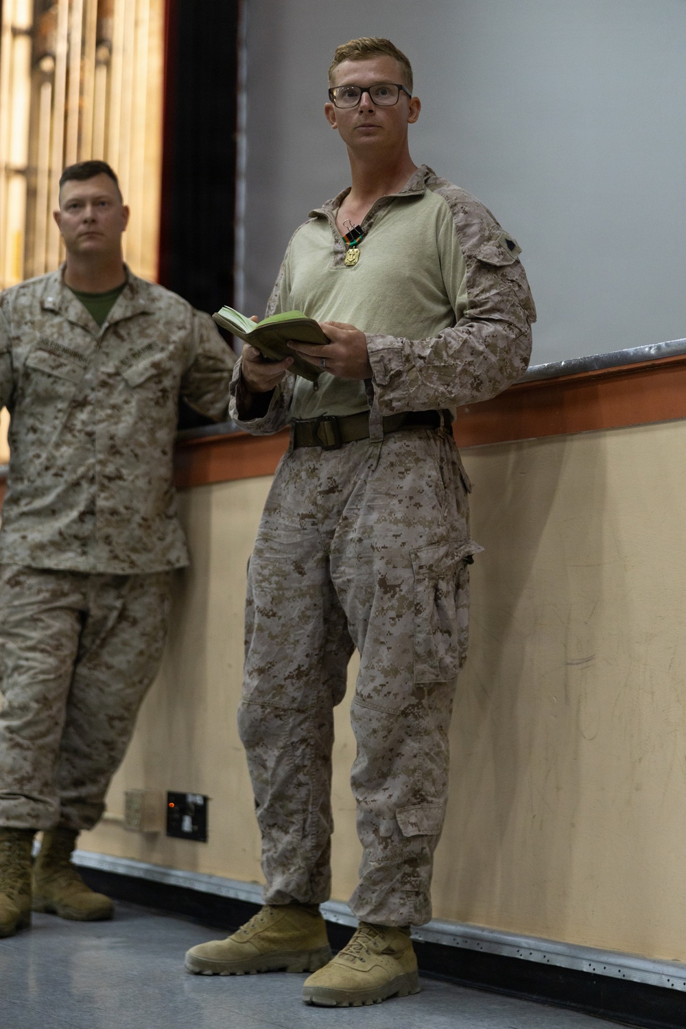 Two military personnel in USMC MARPAT desert camouflage uniforms are standing in what appears to be an indoor briefing or ceremony room. The foreground individual is wearing glasses, holding a small green notebook, and has a medal or award hanging around his neck. A second service member stands slightly behind and to the left.