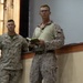 Two military personnel in USMC MARPAT desert camouflage uniforms are standing in what appears to be an indoor briefing or ceremony room. The foreground individual is wearing glasses, holding a small green notebook, and has a medal or award hanging around his neck. A second service member stands slightly behind and to the left.