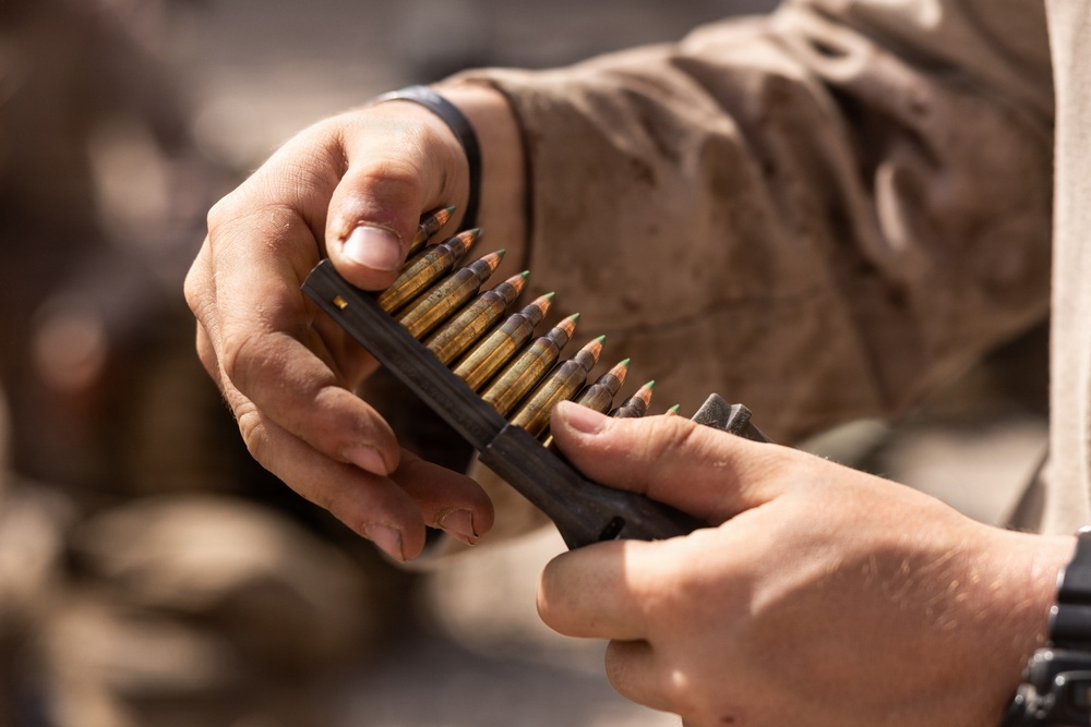 A close-up image showing two hands handling a stripper clip loaded with rifle cartridges featuring green-tipped bullets. The person is wearing desert camouflage military uniform and a black wristband/watch. The image appears to depict a military or tactical ammunition loading scenario.
