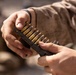 A close-up image showing two hands handling a stripper clip loaded with rifle cartridges featuring green-tipped bullets. The person is wearing desert camouflage military uniform and a black wristband/watch. The image appears to depict a military or tactical ammunition loading scenario.