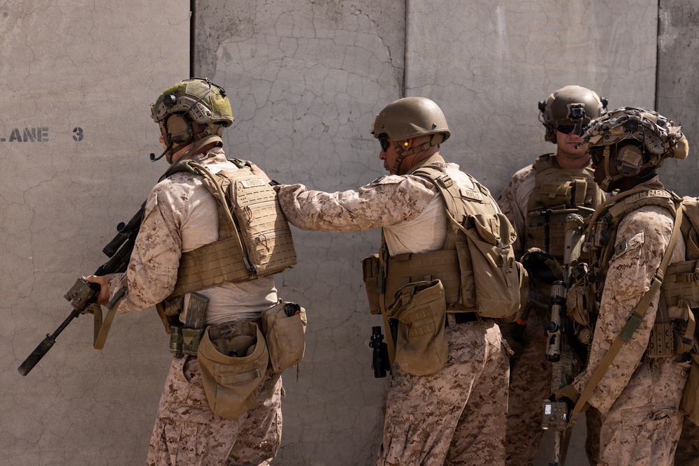 Four military personnel in desert camouflage uniforms are positioned against a concrete wall marked 'LANE 3', appearing to be conducting a tactical training exercise. The soldiers are fully equipped with body armor, helmets, tactical gear, and firearms. One soldier is placing a hand on another's back, suggesting coordination or a 'stack' formation commonly used in close-quarters battle training.