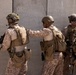 Four military personnel in desert camouflage uniforms are positioned against a concrete wall marked 'LANE 3', appearing to be conducting a tactical training exercise. The soldiers are fully equipped with body armor, helmets, tactical gear, and firearms. One soldier is placing a hand on another's back, suggesting coordination or a 'stack' formation commonly used in close-quarters battle training.