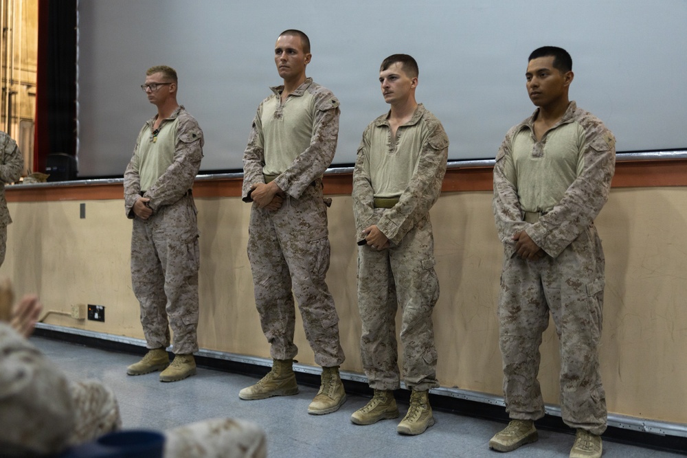 Four military personnel in U.S. Marine Corps MARPAT desert camouflage uniforms stand in a line at what appears to be an indoor ceremony or formation. They are standing at ease with hands clasped in front of them, facing an audience. The setting appears to be a briefing room or auditorium with a projection screen visible in the background.