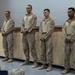 Four military personnel in U.S. Marine Corps MARPAT desert camouflage uniforms stand in a line at what appears to be an indoor ceremony or formation. They are standing at ease with hands clasped in front of them, facing an audience. The setting appears to be a briefing room or auditorium with a projection screen visible in the background.