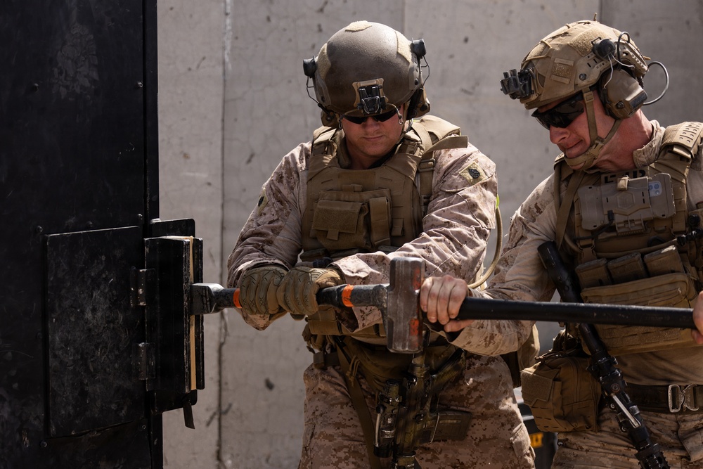 Two military personnel in desert camouflage uniforms are using a breaching tool (sledgehammer/ram) to force open a heavy black metal door. Both individuals are wearing full tactical gear including helmets with NVG mounts, plate carriers, tactical gloves, and sunglasses. The setting appears to be a training or operational environment with a concrete wall backdrop.
