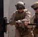 Two military personnel in desert camouflage uniforms are using a breaching tool (sledgehammer/ram) to force open a heavy black metal door. Both individuals are wearing full tactical gear including helmets with NVG mounts, plate carriers, tactical gloves, and sunglasses. The setting appears to be a training or operational environment with a concrete wall backdrop.