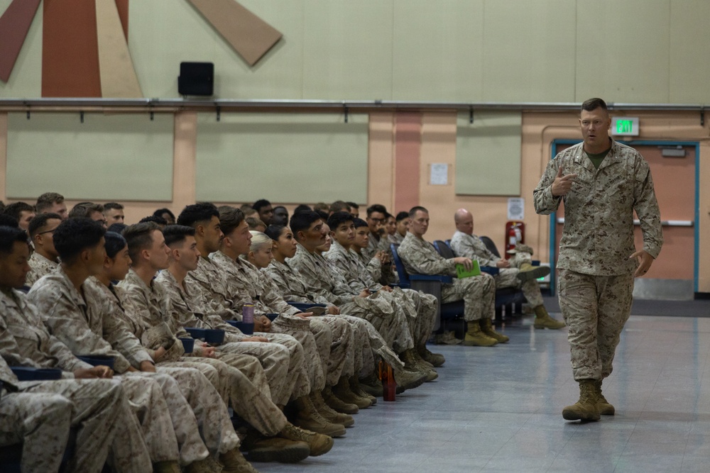 A senior military officer in U.S. Marine Corps MARPAT camouflage uniform addresses a large group of seated Marines in what appears to be a briefing or all-hands meeting inside a military facility. The standing officer appears to be speaking and gesturing while the assembled Marines listen attentively from chairs arranged in rows. The setting suggests a formal command briefing or training session.