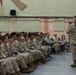 A senior military officer in U.S. Marine Corps MARPAT camouflage uniform addresses a large group of seated Marines in what appears to be a briefing or all-hands meeting inside a military facility. The standing officer appears to be speaking and gesturing while the assembled Marines listen attentively from chairs arranged in rows. The setting suggests a formal command briefing or training session.