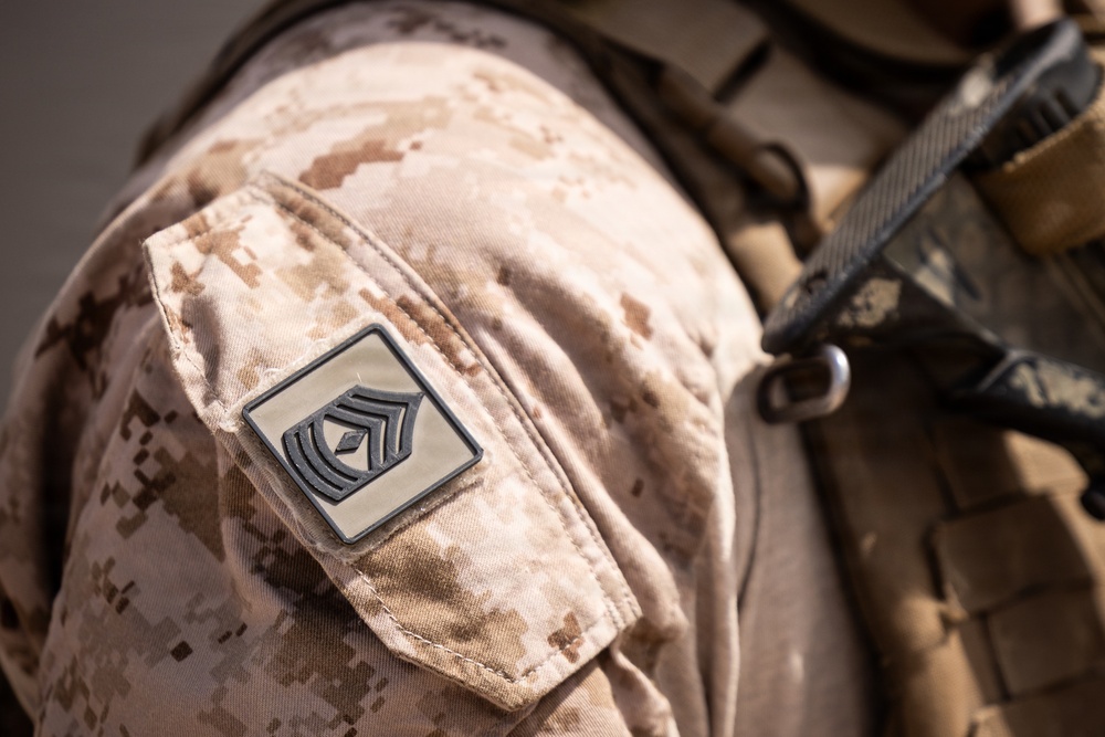 Close-up photograph of a military uniform sleeve displaying a rank insignia patch on desert digital camouflage (MARPAT Desert pattern). The patch shows what appears to be a First Sergeant or Master Sergeant rank insignia with chevrons and a diamond/lozenge symbol. Tactical gear straps and equipment are visible in the background over the right shoulder.