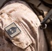 Close-up photograph of a military uniform sleeve displaying a rank insignia patch on desert digital camouflage (MARPAT Desert pattern). The patch shows what appears to be a First Sergeant or Master Sergeant rank insignia with chevrons and a diamond/lozenge symbol. Tactical gear straps and equipment are visible in the background over the right shoulder.